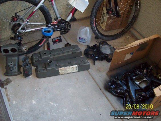 engine parts, including valve covers and intake manifold, laid out on concrete next to two bicycles