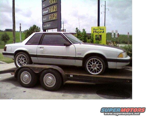 White coupe with black hood on a trailer, side view. Has aftermarket wheels