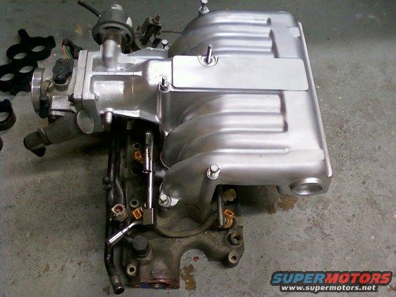 Engine intake manifold with fuel injectors and sensors attached. It is silver and sits on a concrete floor
