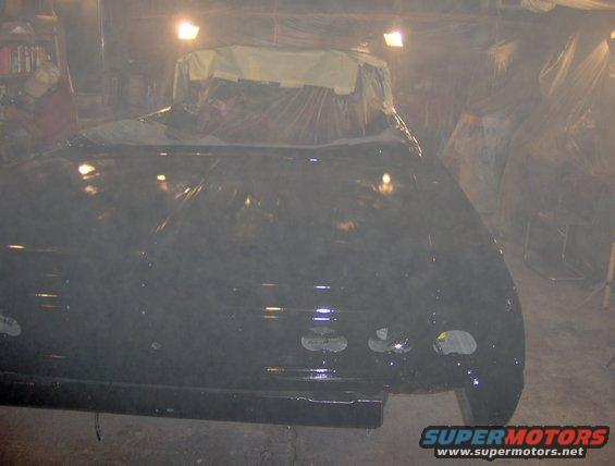 Black car body, possibly a convertible, covered in plastic in a garage with lights on