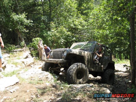 Mud-covered 2005 Jeep Wrangler driving on a rocky, wooded trail