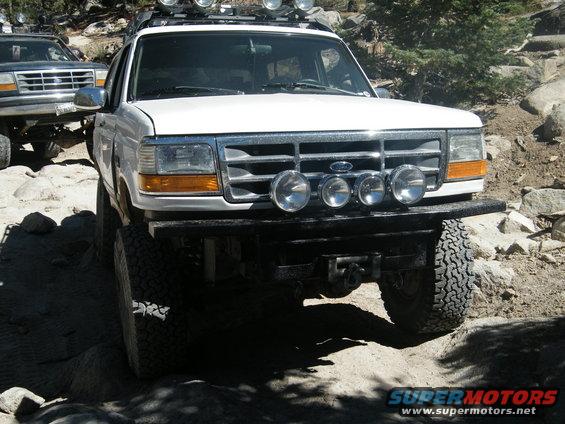 White Ford Bronco with aftermarket bumper and lights, driving on a rocky trail. Another vehicle is visible behind it