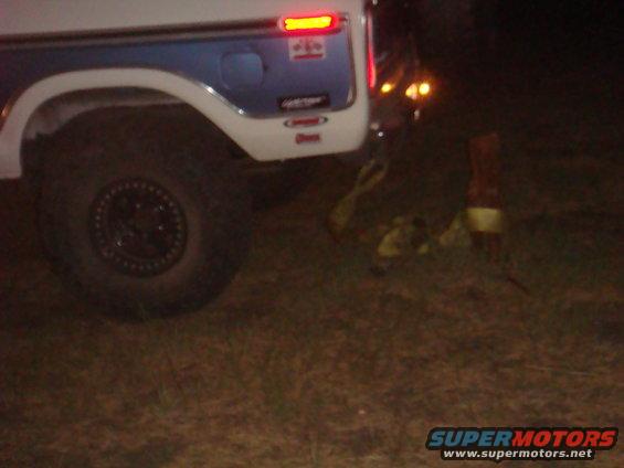 Rear quarter of a 1978 Ford Bronco with a blue stripe and a spare tire mounted on the rear door. Straps are on the ground