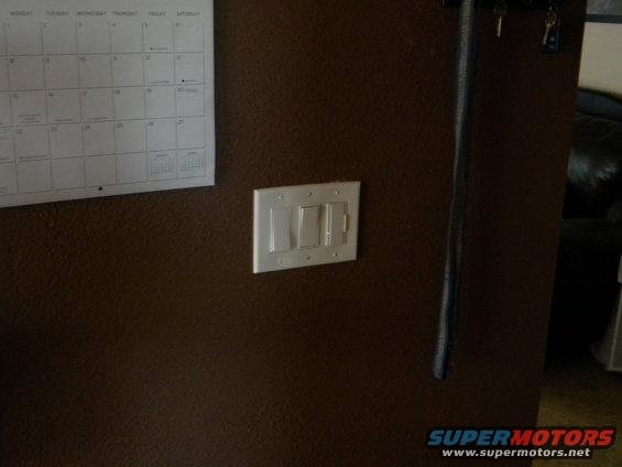 Light switches on a brown wall next to a calendar. A dark object hangs vertically on the right