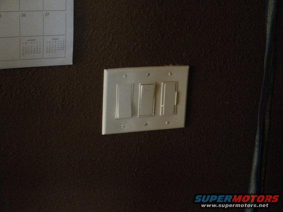 Light switches on a dark wall next to a calendar. One switch has a yellow indicator light