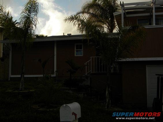 House with palm trees, a white mailbox in the foreground, and a garage door on the right. Supermotors.net logo in the bottom right