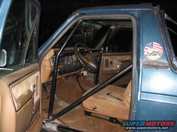 Interior of a blue SUV with a roll cage, tan seats, and a steering wheel. A sticker with an American flag and a truck is on the door