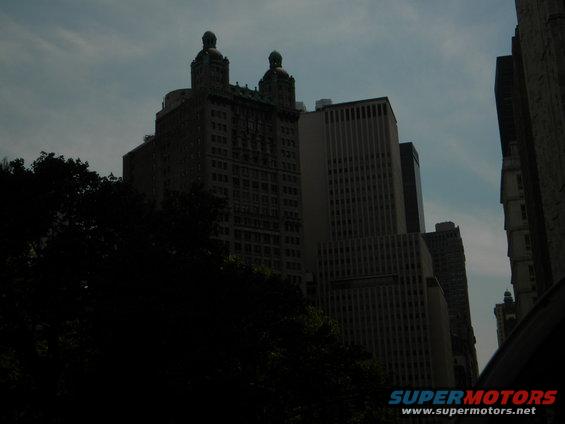 Cityscape with tall buildings and trees under a cloudy sky. Supermotors logo visible