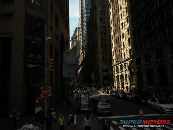 City street with tall buildings, parked cars, and pedestrians. A banner on a building reads '75 WALL'