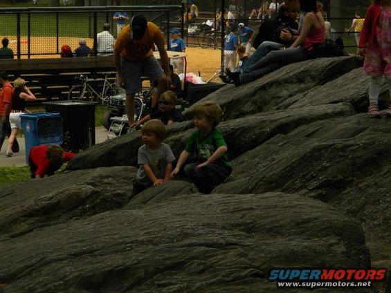 Two young boys sit on large rocks, with a baseball field and spectators in the background
