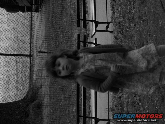 Child in dress and cardigan stands near a chain-link fence and benches