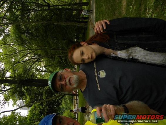 People in a park with trees and grass, one man wears a green cap, another a blue cap, and a woman rests her head on a man's shoulder