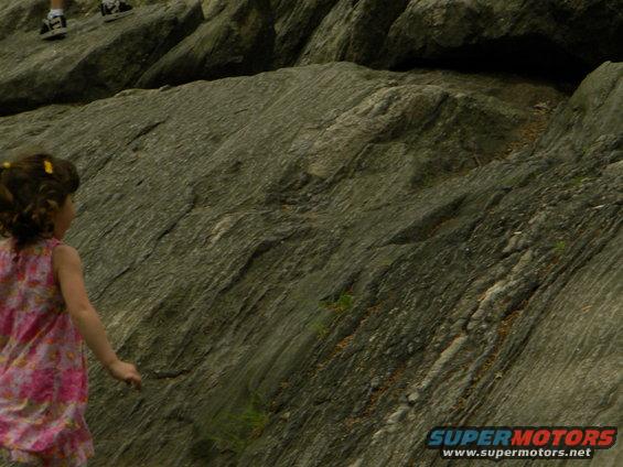 Girl in pink dress looks at rocky terrain. Two people's legs are visible at the top