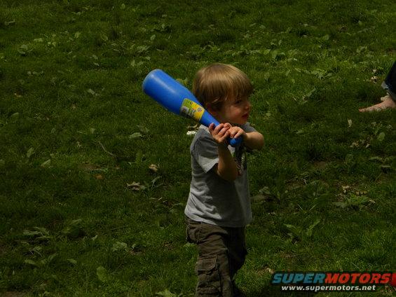 Child in gray shirt and cargo pants holds a blue toy bat in a grassy area