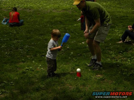 Child swings blue bat at tee-ball, adult watches