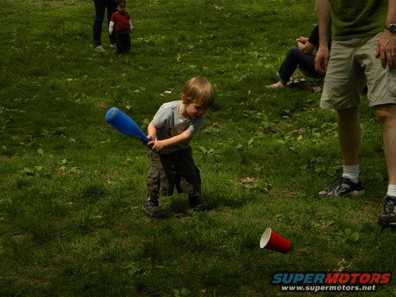 Child with blue plastic bat swings at red cup on grass, with adults and another child in background