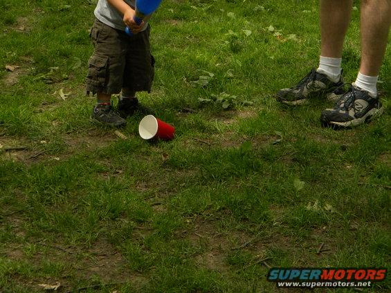 Child in cargo shorts and adult in sneakers stand on grass near a red cup