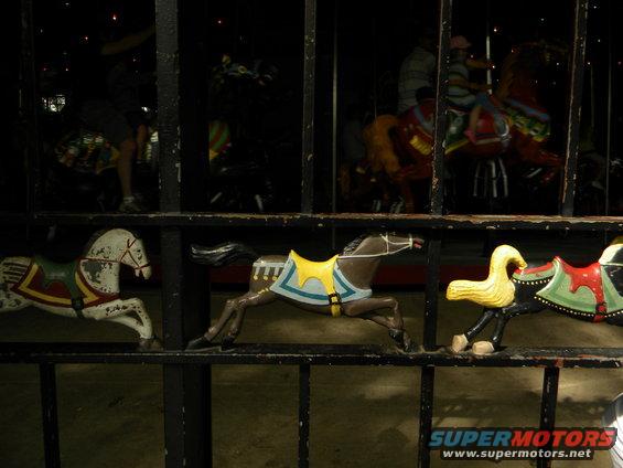 Carousel horses with painted saddles and decorative elements, viewed through a dark metal fence
