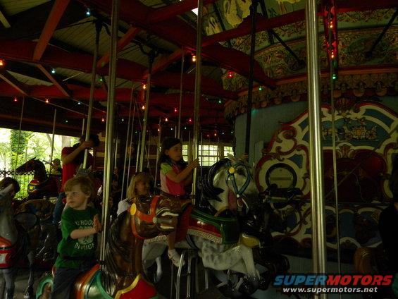 Children ride horses on a carousel, with decorative elements and lights visible