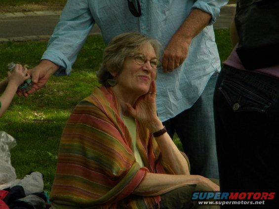 Woman with glasses and a striped shawl sits outdoors, with people behind her