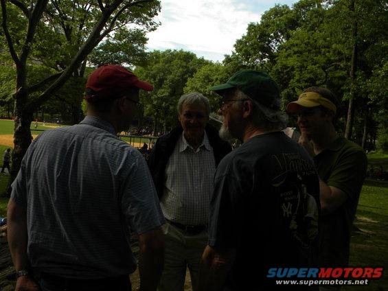 Four men talking in a park with trees and a baseball field in the background. One man wears a red cap, another a green cap, and a third a yellow visor