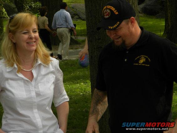 Woman in white shirt and pearl necklace smiles; man in black polo and veteran hat looks down. People in background
