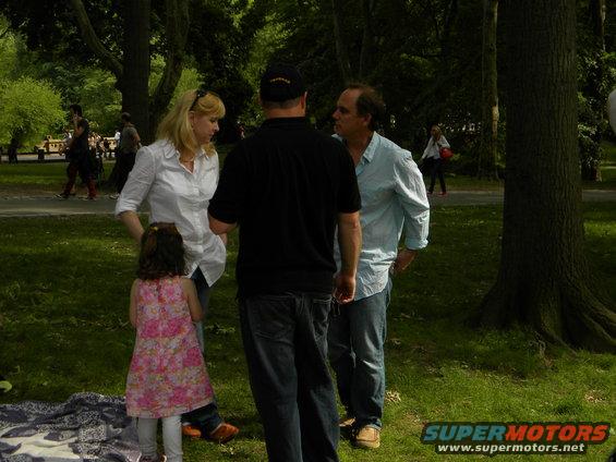 People talking in a park with trees and grass, a woman in a white shirt, a man in a blue shirt, and a child in a pink dress