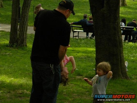 Man holding a child upside down in a park, another child stands nearby