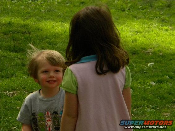 Young boy smiles at the camera while a girl stands with her back to the viewer in a grassy outdoor setting
