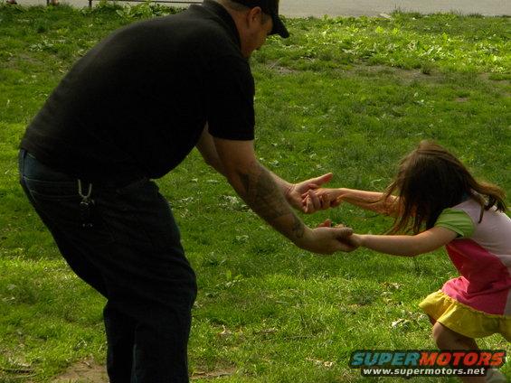Man in black shirt and jeans holding hands with a young girl in a pink and yellow dress, playing in a grassy area