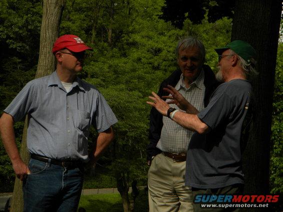 Three men talking outdoors, one wearing a red cap with an 'H'