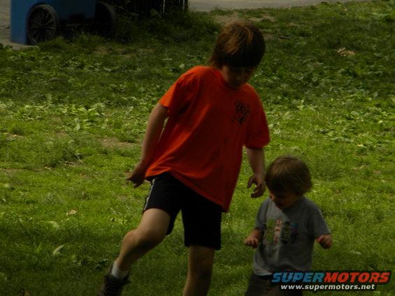 Two young boys are in a grassy area. One boy in an orange shirt kicks his leg up