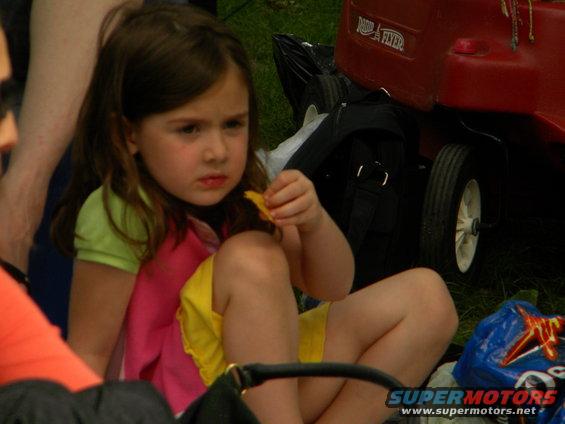 Girl in colorful shirt and yellow shorts sits on grass, holding a chip. A red Radio Flyer wagon is behind her