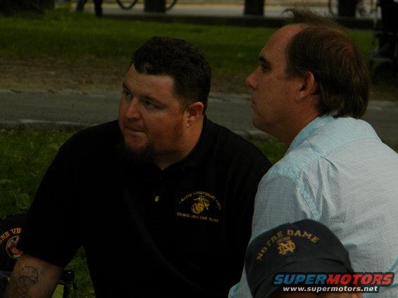 Two men are talking outdoors. One wears a black polo with a Marine Corps emblem. The other wears a light blue shirt. A Notre Dame hat is in the foreground