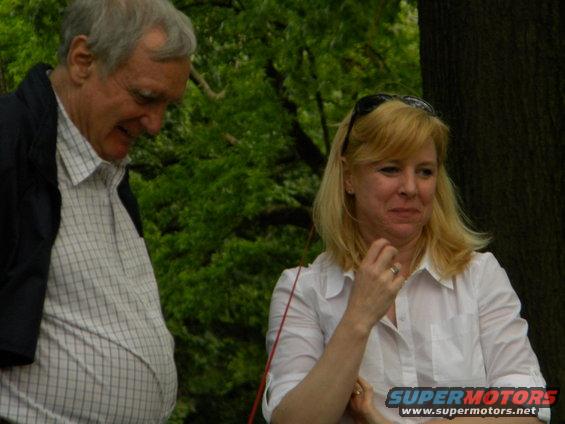 Man in checkered shirt and woman in white shirt outdoors, with a Supermotors logo visible