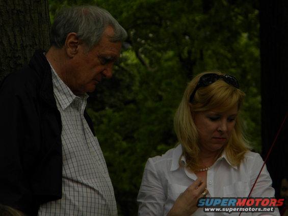 Man and woman looking down outdoors, with trees in the background. A watermark reads SUPERMOTORS.NET