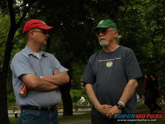 Two men talking in a park, one wearing a red cap, the other a green cap