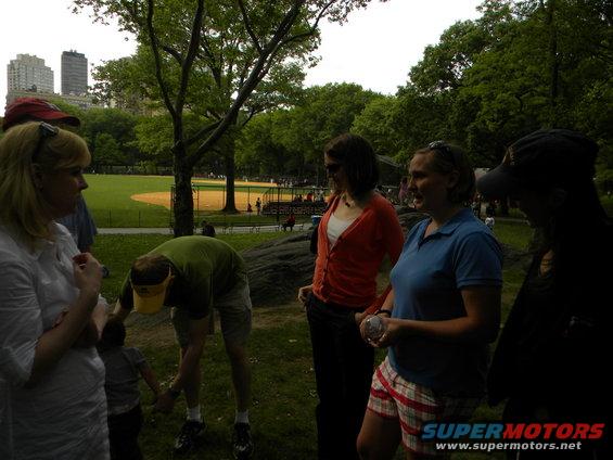 Group of people gathered in a park, with a baseball field and city buildings in the distance