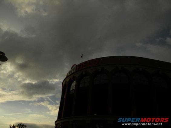 Exterior of a stadium with "FIELD" visible on top, under a cloudy sky. Watermark says SUPERMOTORS