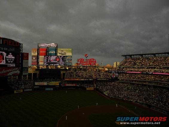 Baseball stadium with filled stands and large scoreboards under a cloudy sky