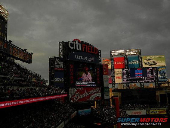 Citi Field stadium exterior with multiple large screens displaying ads and game information under a cloudy sky