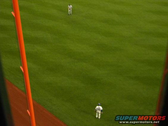 Baseball field with two players in uniform, one in the distance and one closer, with an orange pole in the foreground