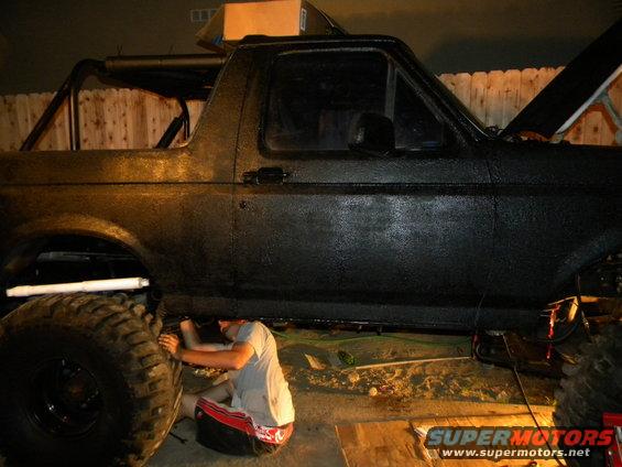Black Ford Bronco with large tires, someone working underneath it. Hood is open