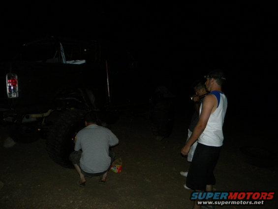 People are gathered around a large, dark off-road vehicle at night. One person is crouching near the rear tire