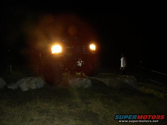 1992 Ford Bronco with headlights on at night, parked on grassy terrain with rocks