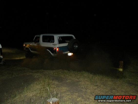 White Ford Bronco with spare tire on back driving on dirt road at night, kicking up dust. Another vehicle is partially visible on the left