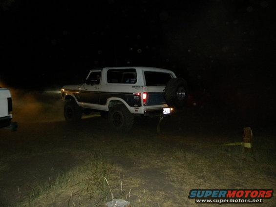 White and blue Ford Bronco parked at night with a spare tire on the back. Another vehicle is partially visible on the left