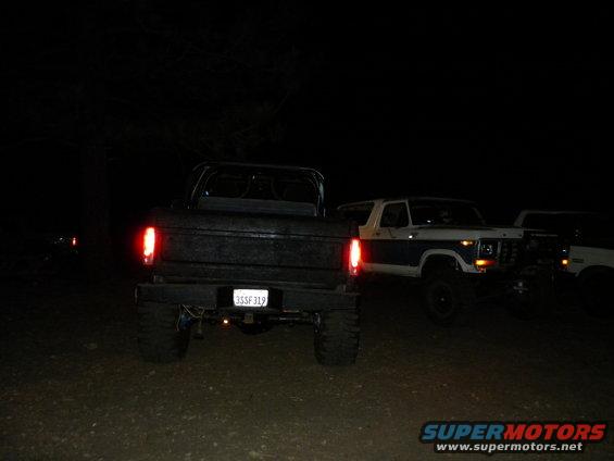 Rear view of a black pickup truck with a white Bronco and another white vehicle behind it at night. License plate reads 3SSF319