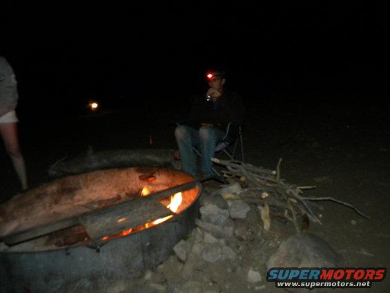 People gathered around a campfire at night, one person wearing a headlamp