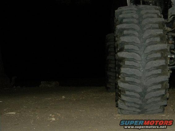 Tire tread of a large vehicle, close-up, with another tire visible behind it in the dark